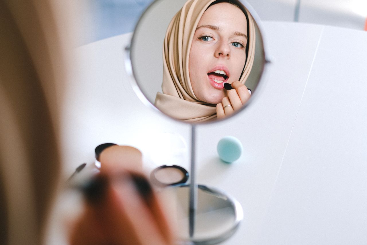 Home Muslim woman in hijab applies lipstick while looking at her reflection in a mirror.