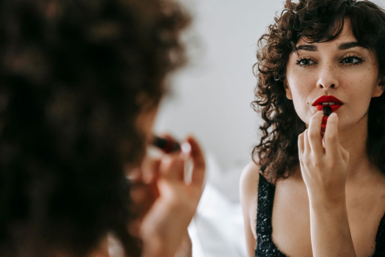 Beautiful female with curly hair applying red lipstick on lips while looking at mirror in light room against blurred background